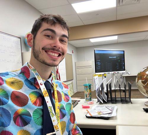 Young male in bright shirt stands in his classroom with a desk, screen, and globe in the background