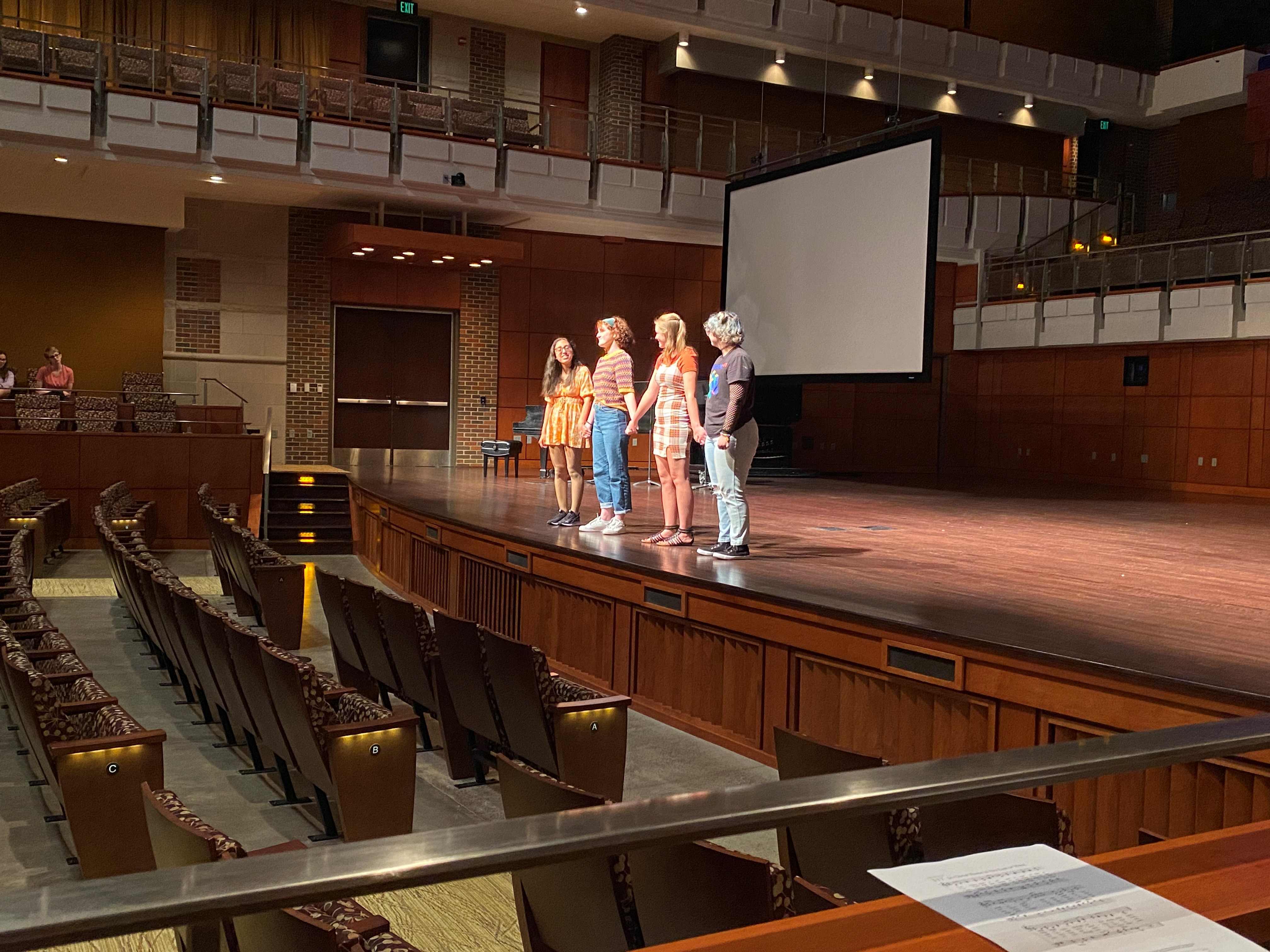 4 students standing on stage in Parmer Hall holding hands while performing a skit for the 2022 National Day of Peace Event