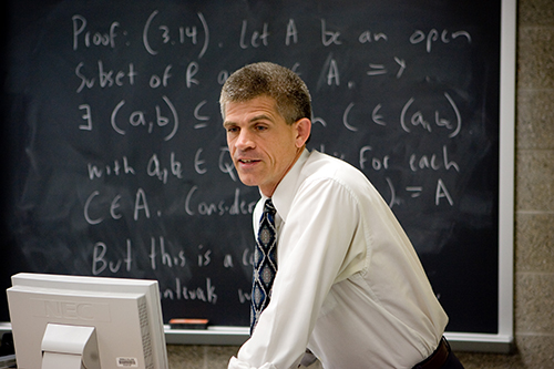A math professor standing in front of a filled black board.