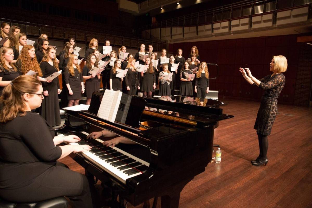 A female conductor and her choir, accompanied by a pianist.