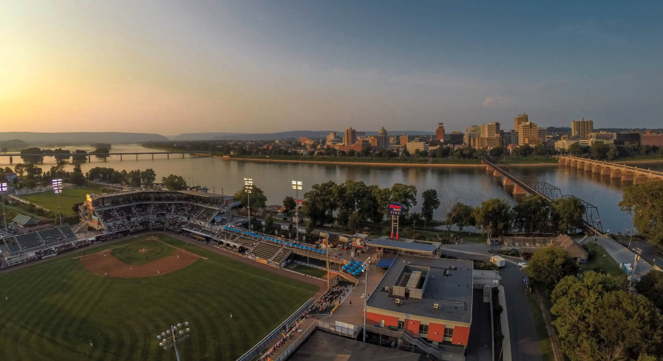 A wide aerial view of a riverside baseball stadium during a game, with the field glowing under stadium lights as players look tiny enough to be action figures. Beyond the outfield, a calm river stretches across the scene, with bridges connecting to a downtown skyline full of tall buildings basking in golden-hour light. The whole landscape gives off strong “postcard you’d buy because the city looks extra photogenic today” energy.
