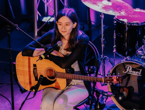 A young musician sits onstage with an acoustic guitar resting across her lap, leaning toward a microphone as purple stage lights make everything look extra magical. A drum set glints behind her like it’s waiting for its big moment. She appears calm and ready to play—kind of like she’s about to drop the most soothing banger in acoustic history.