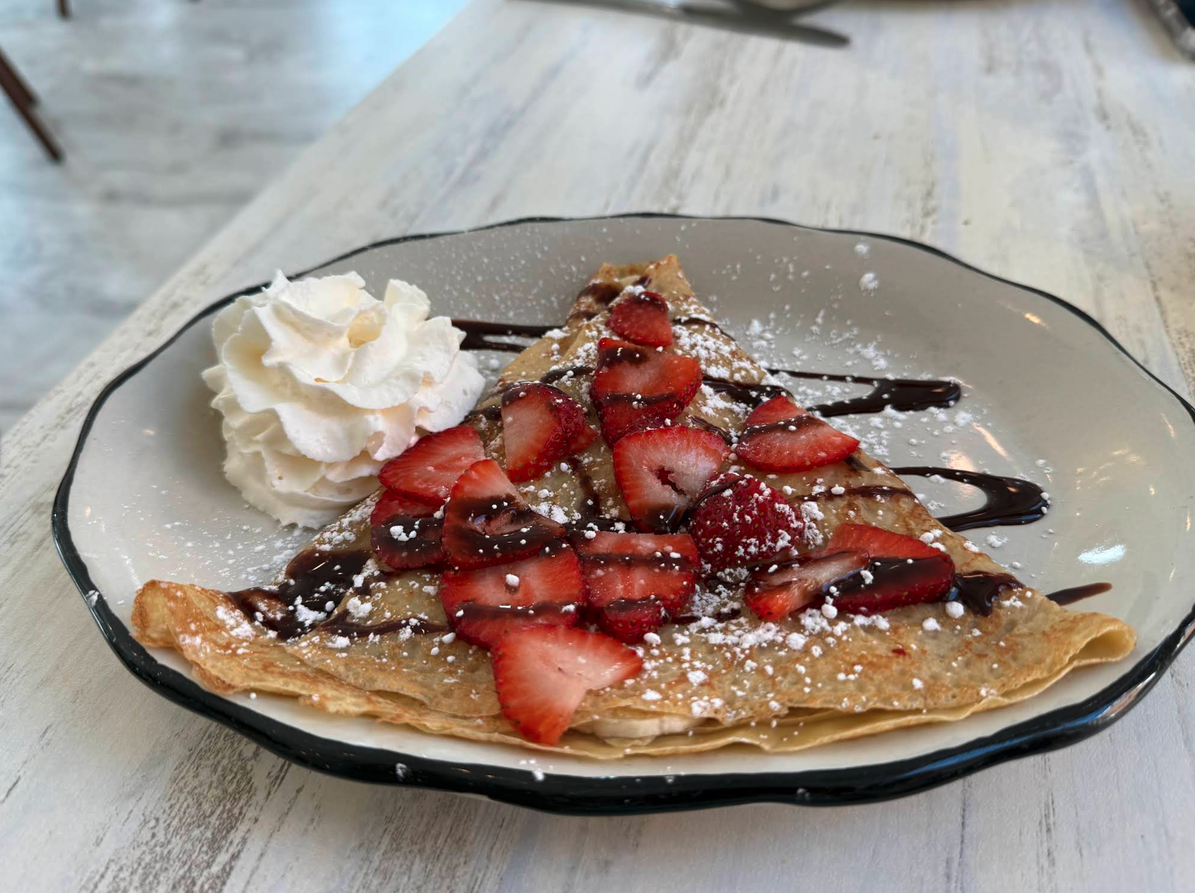 A charming, pastel-colored café decked out with chandeliers, floral garlands, and all the cozy décor a Pinterest board could dream of. Behind the counter, a staff member works at a register surrounded by trays of pastries, tiny French flags, and handwritten signs on the mirror. Shelves display teacups, décor, and Eiffel Tower art, giving the whole place strong “welcome to Paris, but make it extremely adorable” vibes.