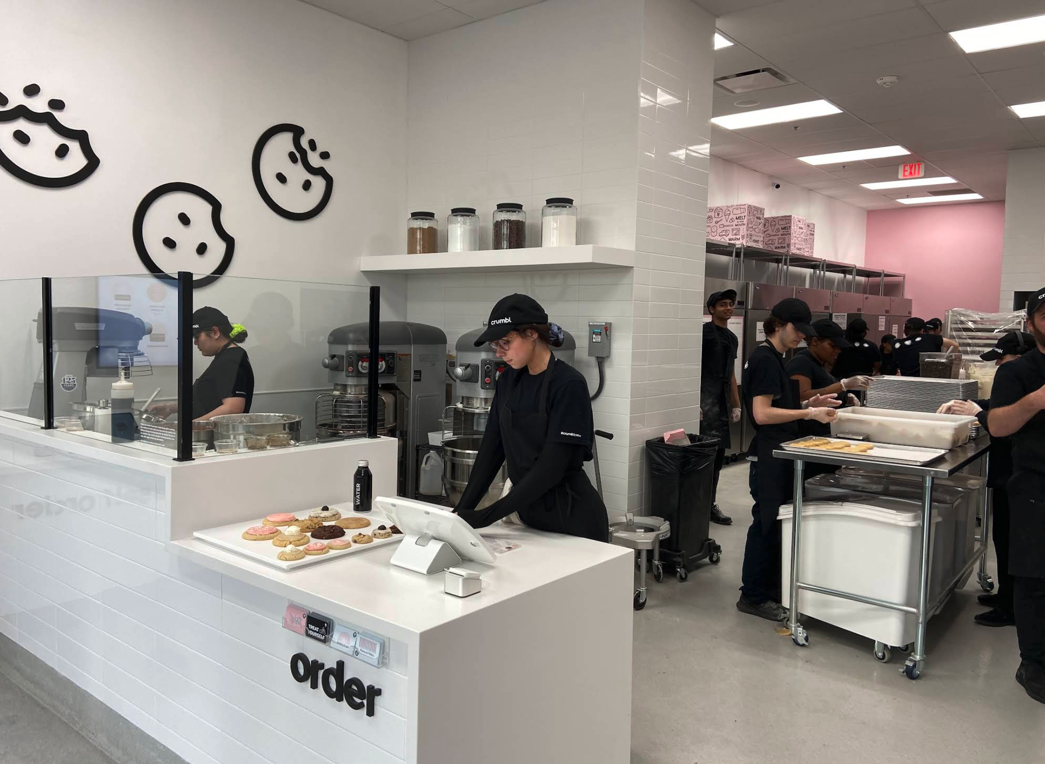 Workers in a bright, modern cookie shop prepare baked goods behind a glass counter while one employee stands at the register beside a tray of perfectly arranged cookies—each one looking like it’s auditioning to be “cookie of the month.” The walls feature cute cookie-face decorations, large mixers hum in the background, and the kitchen bustles with staff assembling ingredients like a highly coordinated dessert-making orchestra.