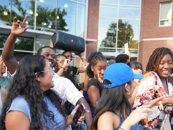 A lively group of students dances and laughs together outdoors, packed closely like they all simultaneously heard their favorite song. One person holds a large speaker over their shoulder—full “human boombox” mode—while others gesture, smile, and groove. Sunlight reflects off nearby windows, adding a festive glow to the already high-energy scene, as if the whole crowd collectively decided, “Yep, this is now a music video.”