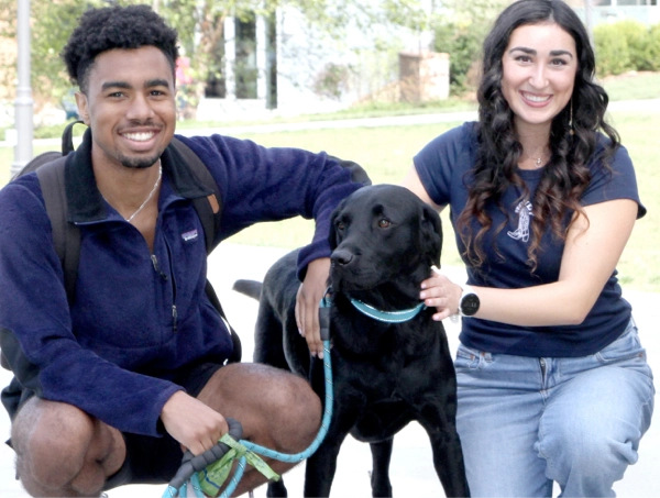Two smiling students crouch on a sidewalk petting a large black dog who looks very pleased with its newfound fan club. One student holds the leash while the other gives the dog a gentle shoulder pat—basically the canine equivalent of a VIP meet-and-greet. Trees and campus buildings blur softly in the background, making the trio look like the cover photo for “Reasons College Is Better With Dogs.”