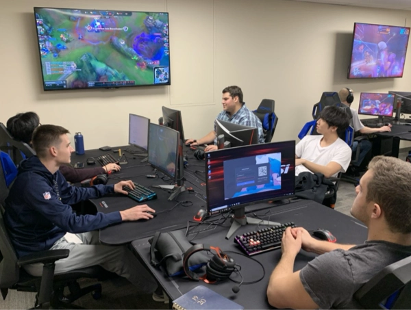A group of students sits at a long table packed with gaming PCs, headsets, and keyboards, fully locked in like they’re competing for the title of “Most Intense Clicking.” Several large screens on the wall display a colorful multiplayer game, and everyone looks focused—except the one guy who seems to be mid–tech troubleshooting, the eternal hero of every gaming session. The room has strong “esports command center” energy, complete with the unspoken agreement that snacks are absolutely allowed.