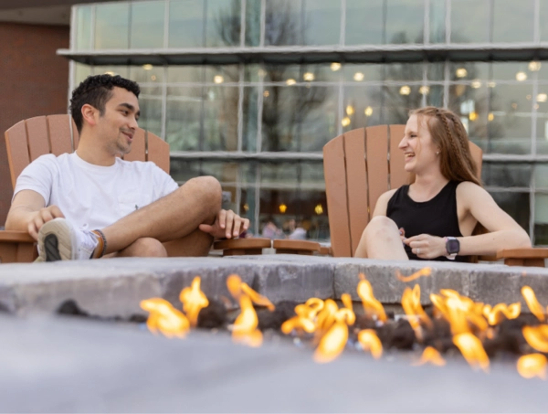 Two students lounge in wooden chairs around an outdoor fire pit, chatting and smiling like they’ve just discovered the secret to eternal relaxation. Soft yellow flames flicker in the foreground, giving the scene major “cozy brochure moment” vibes. A glass-front campus building glows behind them, completing the aesthetic of “homework? never heard of her.”