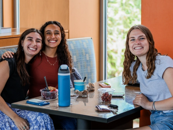Three smiling young women sit around a café table, leaning in like they’re starring in a commercial for friendship. They have bowls of food, fruit cups, water bottles, and phones scattered across the table—classic “we totally meant to study but got distracted by snacks” energy. Bright natural light comes through a window behind them, giving the whole scene cheerful “campus hangout vibes.”