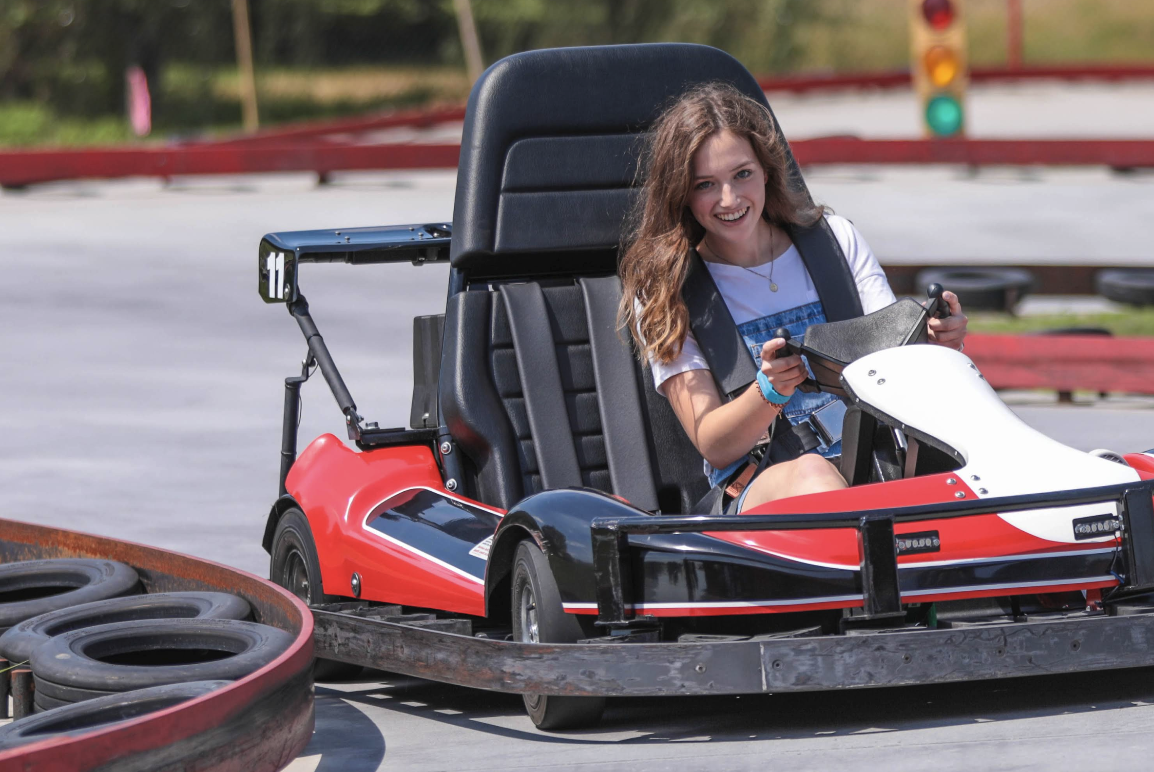 A young person drives a red and black go-kart around a curved track bordered by tires and metal rails. They are seated upright, holding the steering wheel with both hands, and wearing a safety belt. A traffic light in the background shows a green signal, and the track surface and barriers are clearly visible in bright daylight.