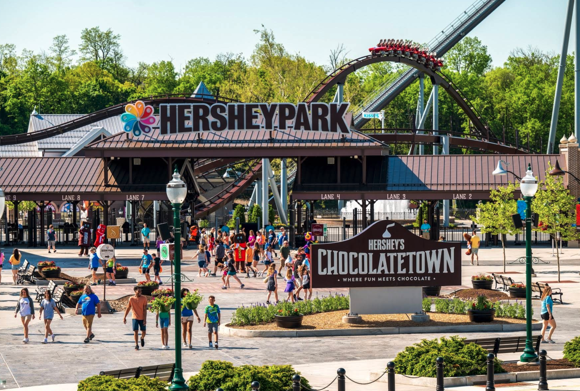 Entrance plaza of Hersheypark on a sunny day, with crowds of visitors walking toward the gates. A large “HERSHEYPARK” sign sits above the entrance lanes, and a roller coaster with a train of riders loops behind it. In the foreground, a sign reading “Hershey’s Chocolatetown—Where Fun Meets Chocolate” stands among landscaped plants and pathways.