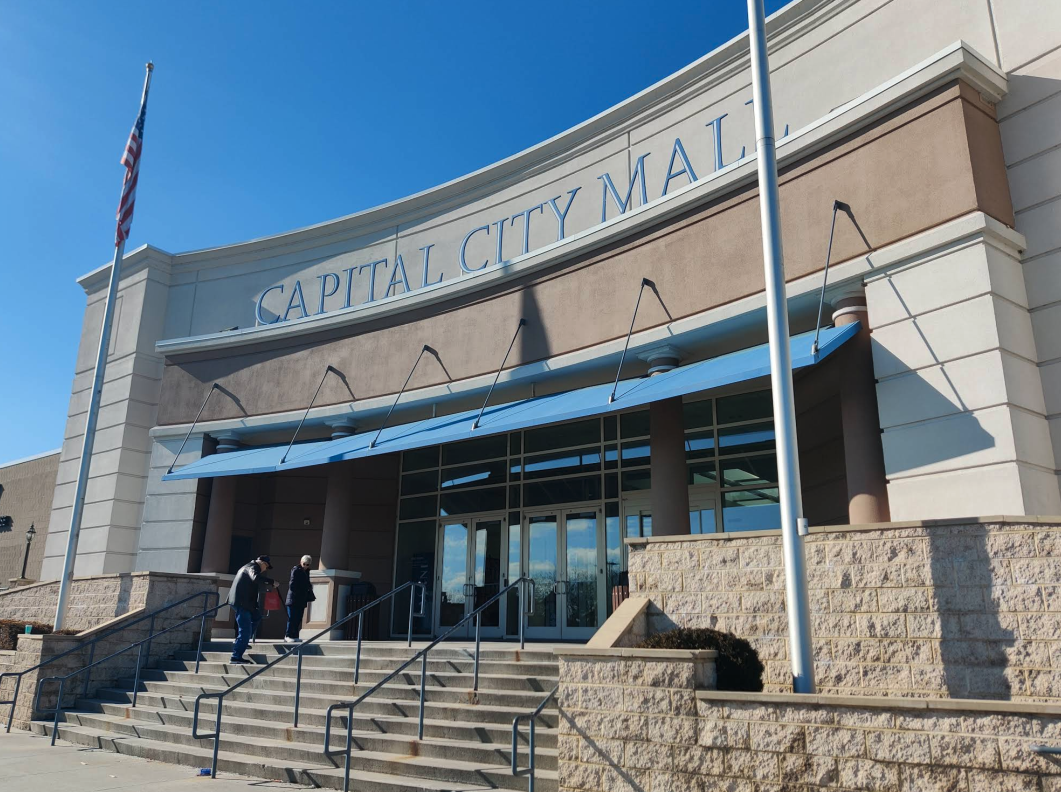 The front entrance of Capital City Mall, featuring a wide staircase leading up to large glass doors beneath blue awnings. A few people walk up the steps, and tall flagpoles frame the scene against a clear blue sky. The building’s curved facade and big block letters give it strong “come inside and maybe buy things you don’t need” energy.