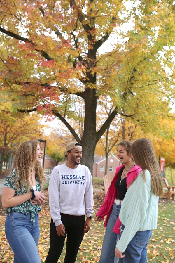Four college students stand outdoors on a campus walkway lined with autumn trees, laughing and talking together. One wears a sweatshirt with “Messiah University” on the front, and the ground around them is covered in fallen leaves.