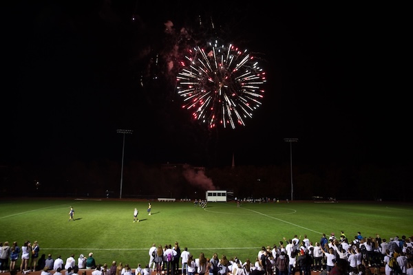 A large crowd of spectators lines the edge of a lit sports field at night while players stand on the grass. Above them, a bright burst of red and white fireworks illuminates the dark sky.