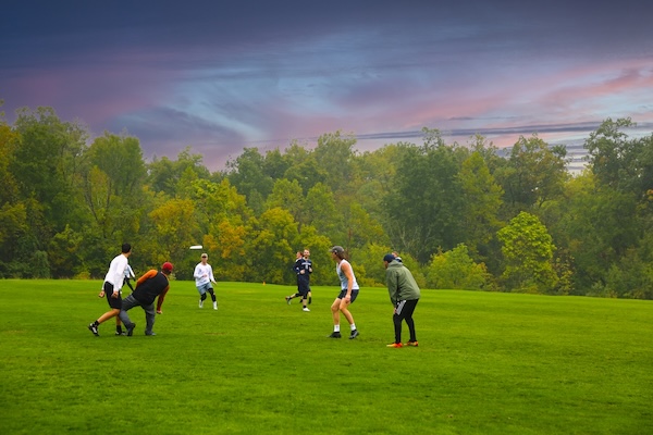 A group of people play an outdoor game of ultimate frisbee on a wide grassy field. One player throws the disc while others run or prepare to catch it, with dense green trees and a colorful, dramatic sky in the background.