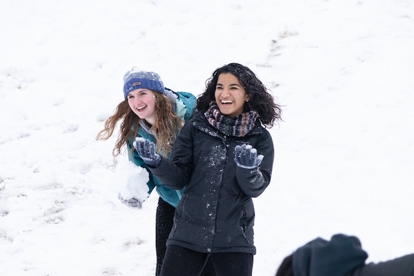Two people stand outdoors in the snow, smiling and mid-throw during a playful snowball fight. Both wear winter coats, gloves, and hats or scarves, with snow covering their clothing and the ground around them.