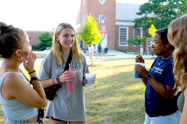 A small group of students stands chatting on a sunny campus lawn. One person holding two drinks smiles while talking, and others around her sip beverages or listen. A brick academic building and trees are visible in the background.