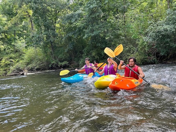 A group of people paddle brightly colored kayaks—orange, yellow, and blue—down a shallow, fast-moving river. They wear life jackets and hold yellow paddles as they navigate the water, with dense green trees lining both sides of the river.