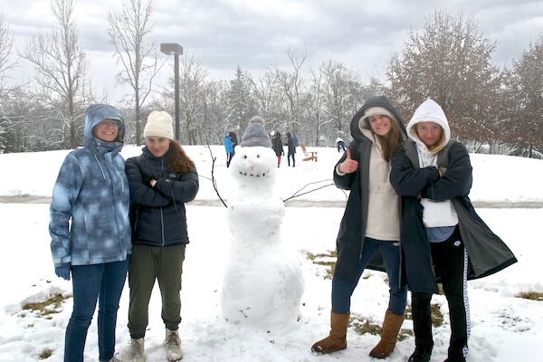 Four people in winter coats and hats stand in the snow posing beside a snowman with stick arms, a gray knit hat, pebble eyes, and a carrot nose. A snowy field with other people playing in the background stretches behind them.