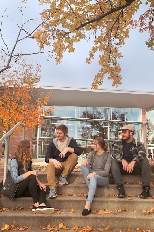 Four people sit talking on an outdoor set of concrete steps covered with fallen autumn leaves. Above them, tree branches with orange and yellow foliage frame the scene. A modern glass-front campus building stands in the background.