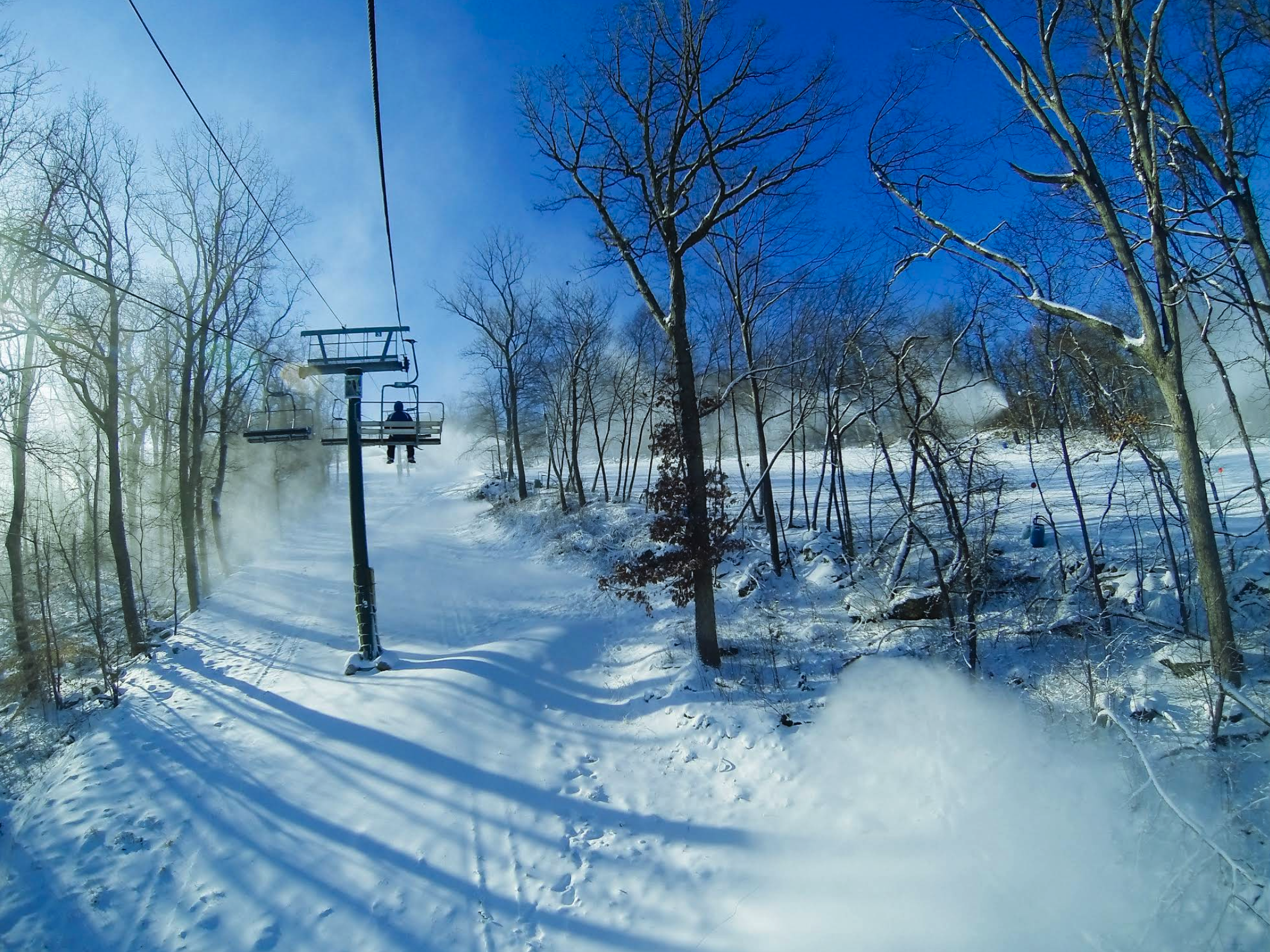 A ski lift carries riders above a snowy slope lined with bare winter trees, all under a bright blue sky so clear it feels like nature’s screensaver.