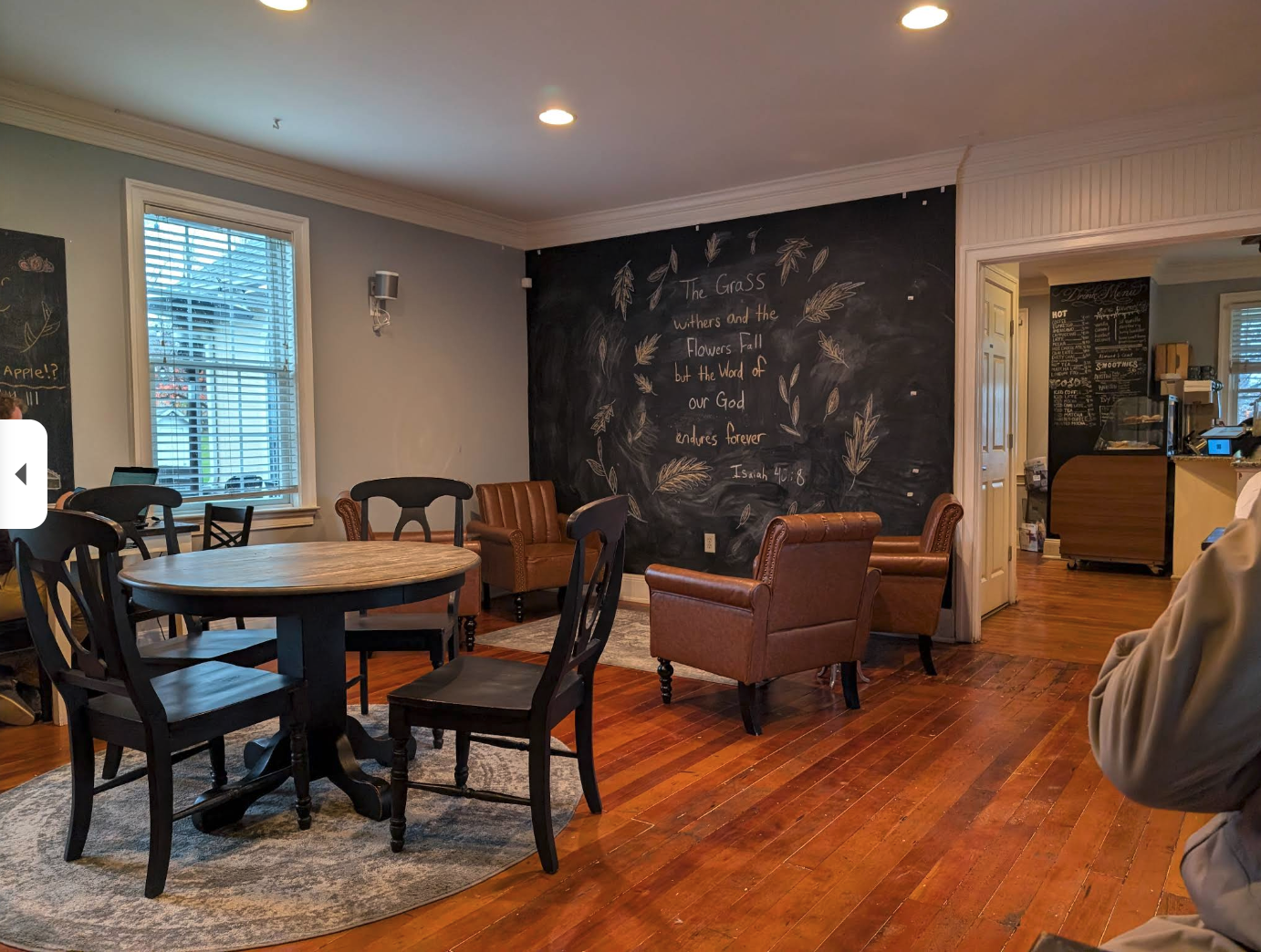 A cozy café-style room with wooden floors, a round table with dark chairs, and a seating area of cushy brown armchairs arranged for maximum “let’s pretend we’re productive” comfort. A large chalkboard wall features a handwritten Bible verse surrounded by leafy doodles, giving the space major artsy-coffeehouse vibes. In the background, a small counter area and menu board peek through a doorway, while part of someone’s shoulder enters the frame like they’re making a dramatic cameo.