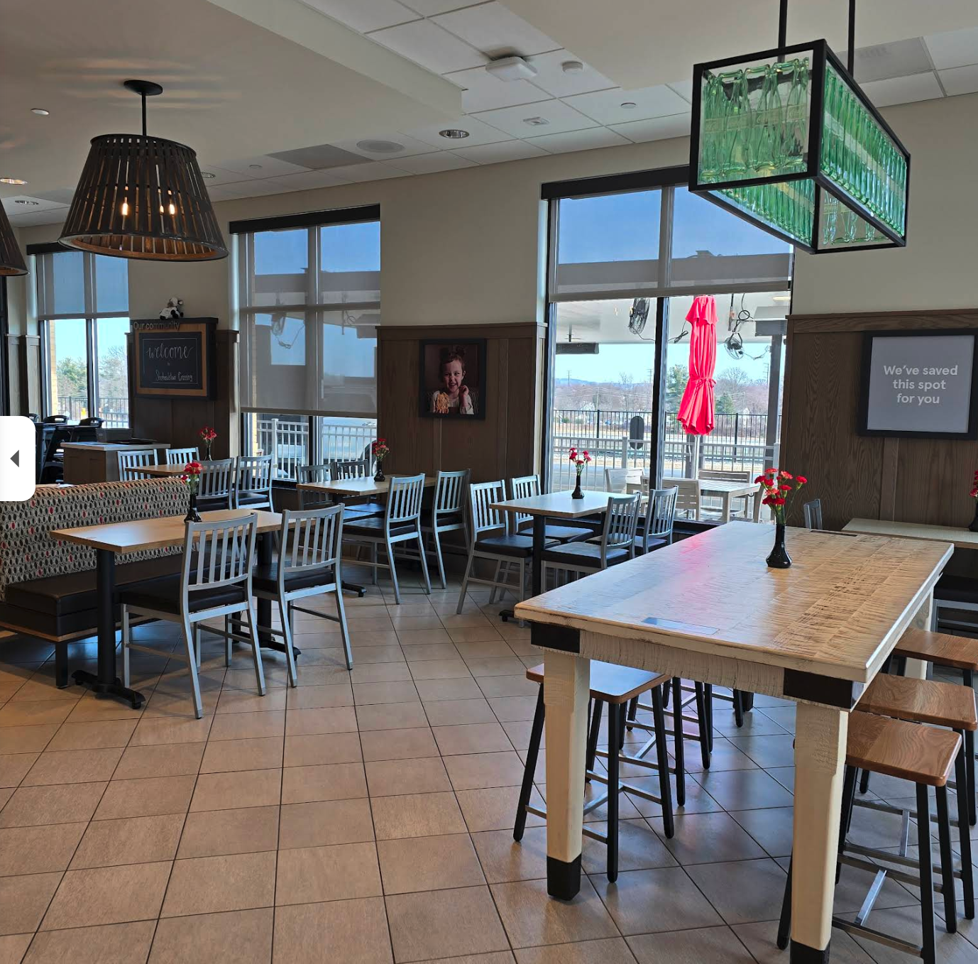 A bright, tidy café-style dining area with several empty tables and chairs arranged neatly across a tiled floor. Small vases with red flowers sit on the tables, adding a touch of “we tried to be fancy” charm. Large windows let in lots of natural light, showing an outdoor patio with red umbrellas.