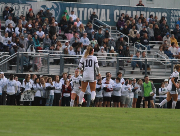 A women’s soccer match in a packed stadium, with a player wearing jersey number 10 standing on the field facing a lively crowd. Teammates and spectators line the sidelines, many in white shirts, while the stands behind them are filled with bundled-up fans. A large banner above the bleachers celebrates past national championships. The whole scene buzzes with energy—like everyone collectively decided, “Yes, today is the perfect day to yell enthusiastically at a ball.”