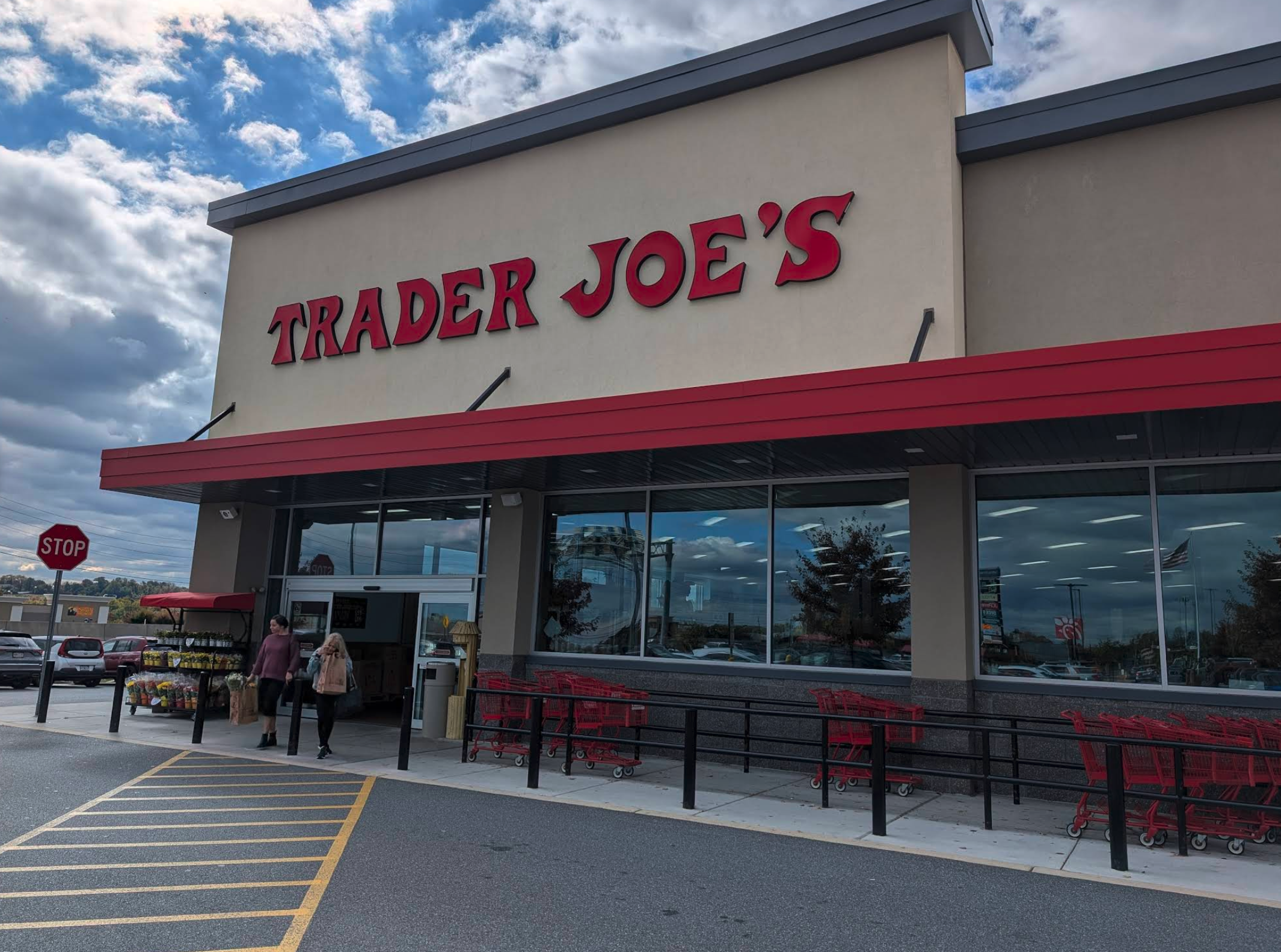The exterior of a Trader Joe’s store with large red lettering and a red awning, set against a partly cloudy sky. A few shoppers walk out with grocery bags while rows of bright red shopping carts wait by the entrance, all giving off the classic “come for one thing, leave with twelve snacks you didn’t plan on” atmosphere.