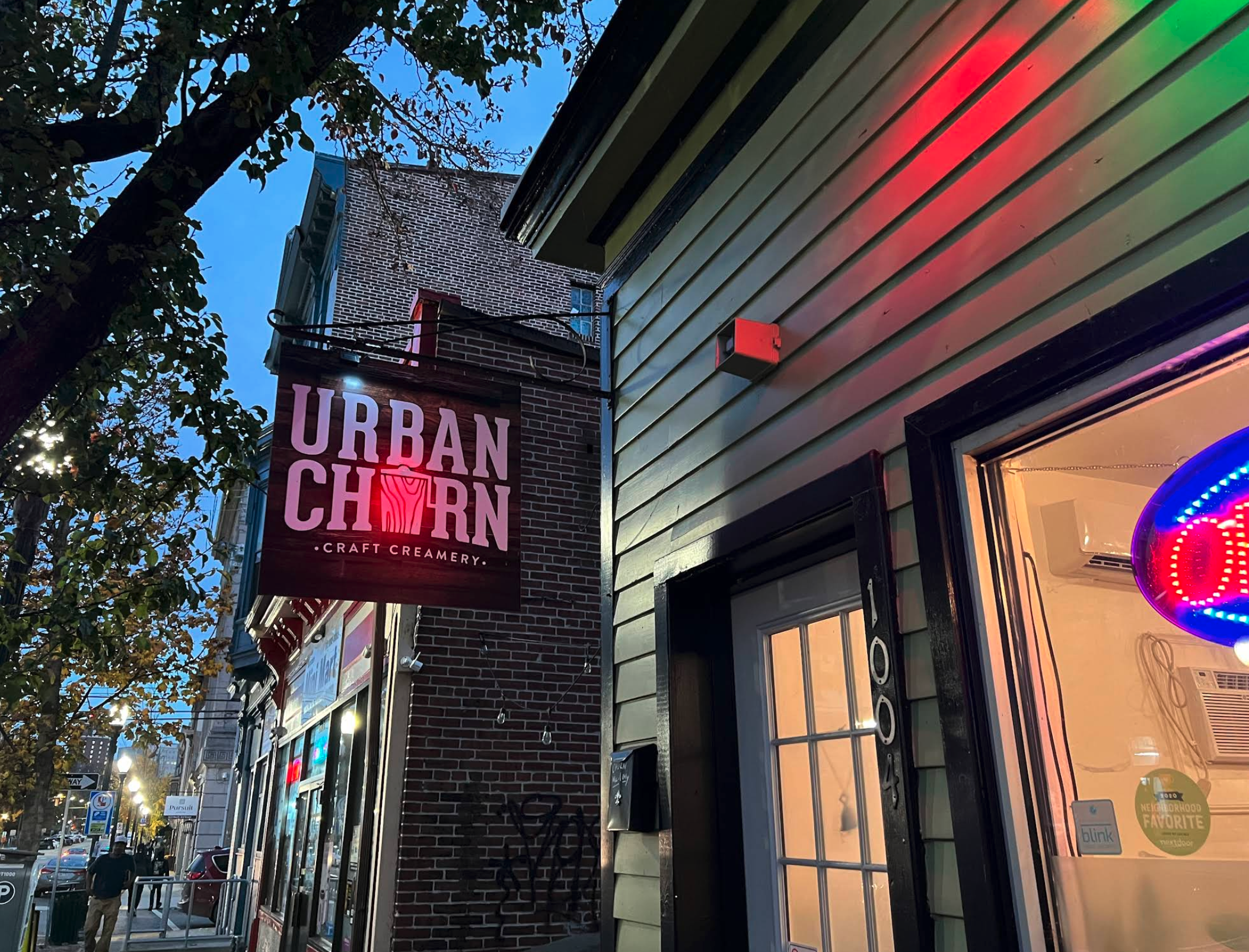 A street-side view of a small storefront at dusk, featuring a lit hanging sign that reads “Urban Churn Craft Creamery.” The building has light-colored siding with dark trim, and a bright “open” sign glows in the front window. Nearby buildings, streetlights, and pedestrians are visible along the sidewalk, with tree branches partially framing the scene.