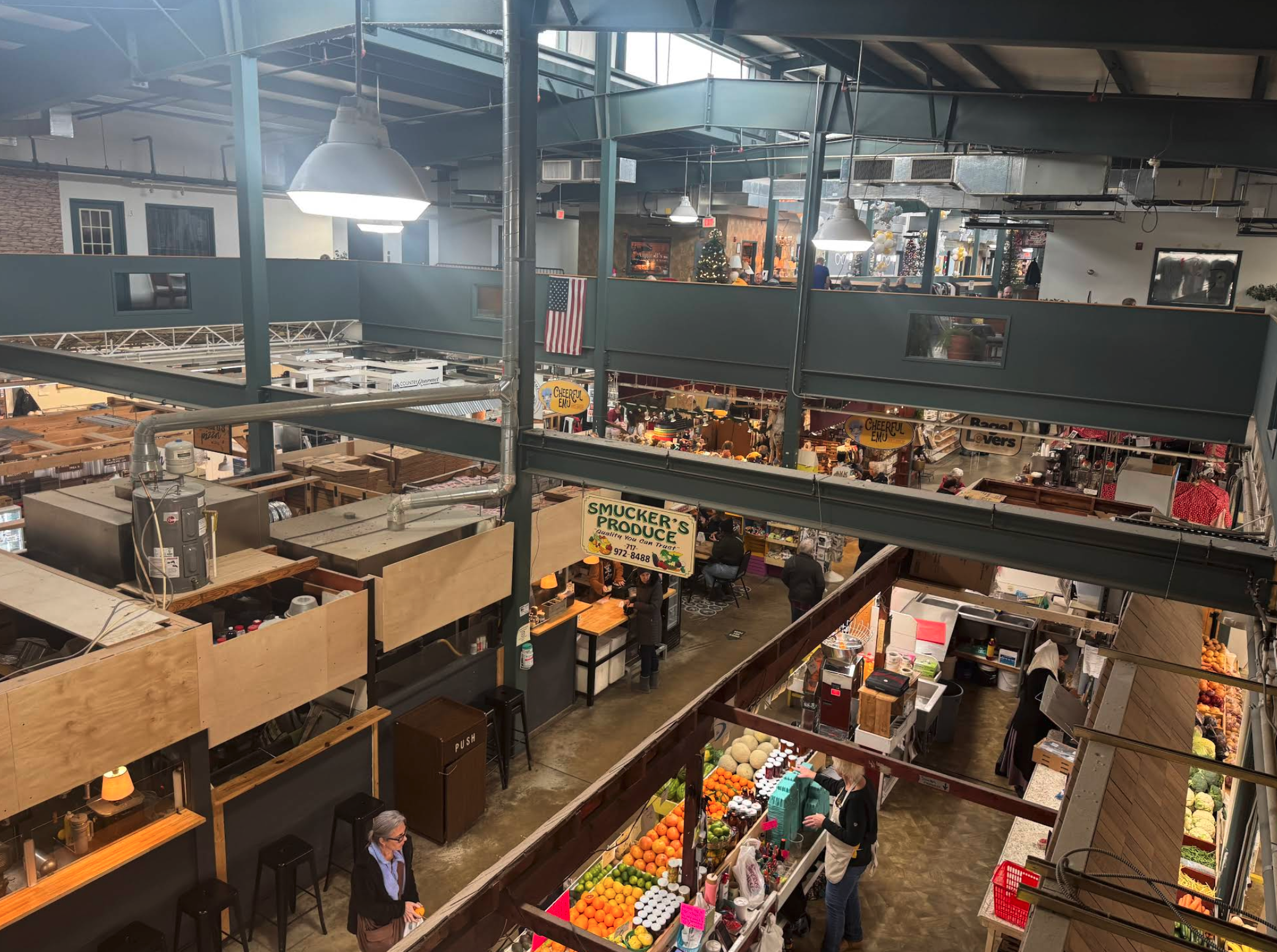 An indoor market viewed from an upper level, showing multiple vendor stalls arranged in rows beneath exposed metal beams and industrial lighting. Produce stands display a variety of fruits and vegetables, while other booths offer prepared foods and goods. Several shoppers and vendors are visible throughout the space. An American flag hangs from the upper level, and signage for different stalls, including “Smucker’s Produce,” is prominently displayed.