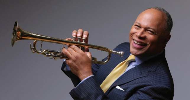 A smiling man in a suit holds a trumpet, showcasing his enthusiasm for music.
