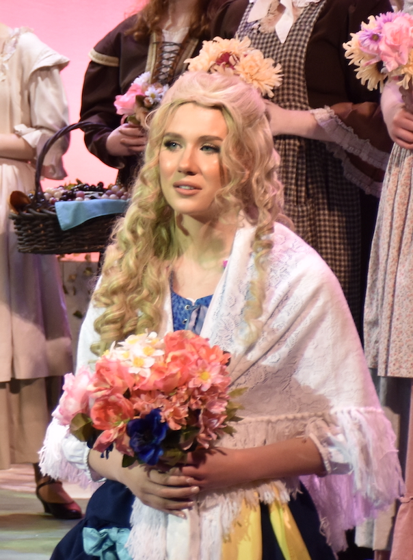 A young actress dressed in period attire holding a bouquet of flowers, with a soft expression while seated in front of an audience backdrop.