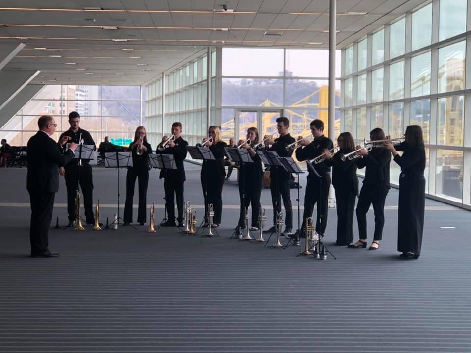 A brass ensemble performs in a spacious, modern venue with large windows showcasing a city view.