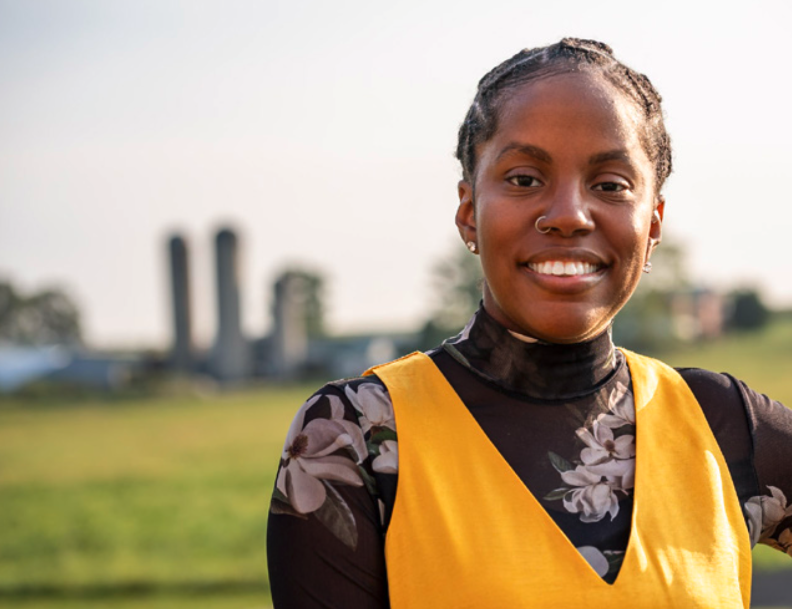 A smiling woman in a yellow dress over a floral top stands against a rural backdrop with silos in the distance.