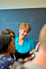 A smiling woman in a blue jacket interacts with individuals in a classroom setting, surrounded by students holding colorful papers.