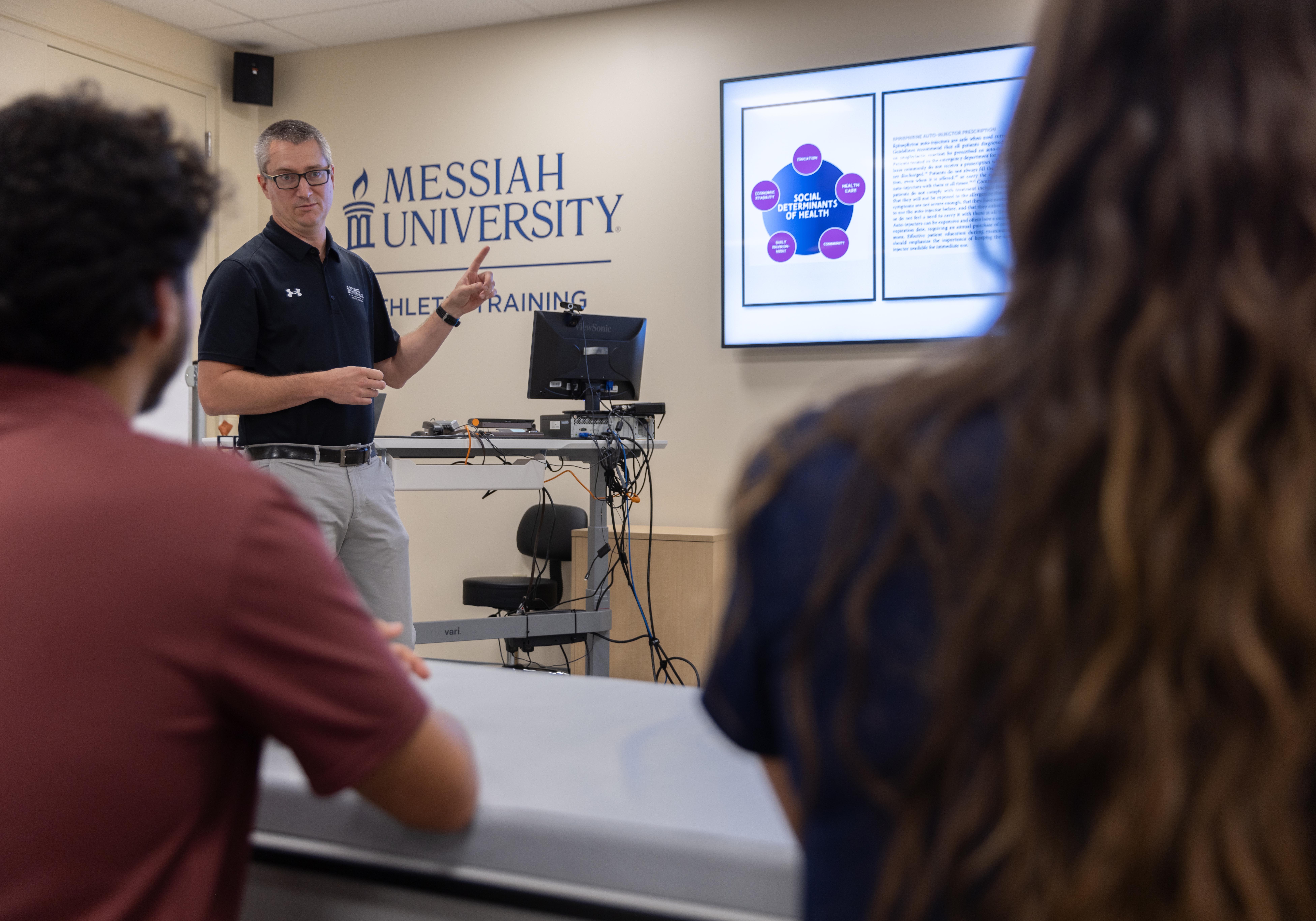 A male teacher is lecturing in front of the class. You can see Messiah University on the board behind him.