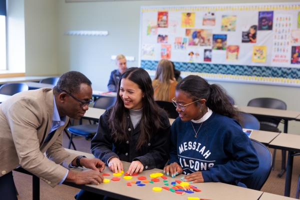 A male professor and two female students sitting at a table look at colorful shape manipulatives together.