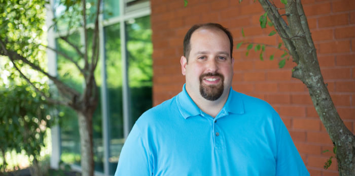 Professor in a turquoise shirt stands in front of a brick wall.