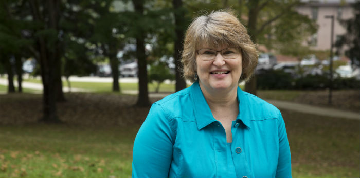 Female stands outside, wearing a turquoise shirt. She is looking straight at the camera, smiling.