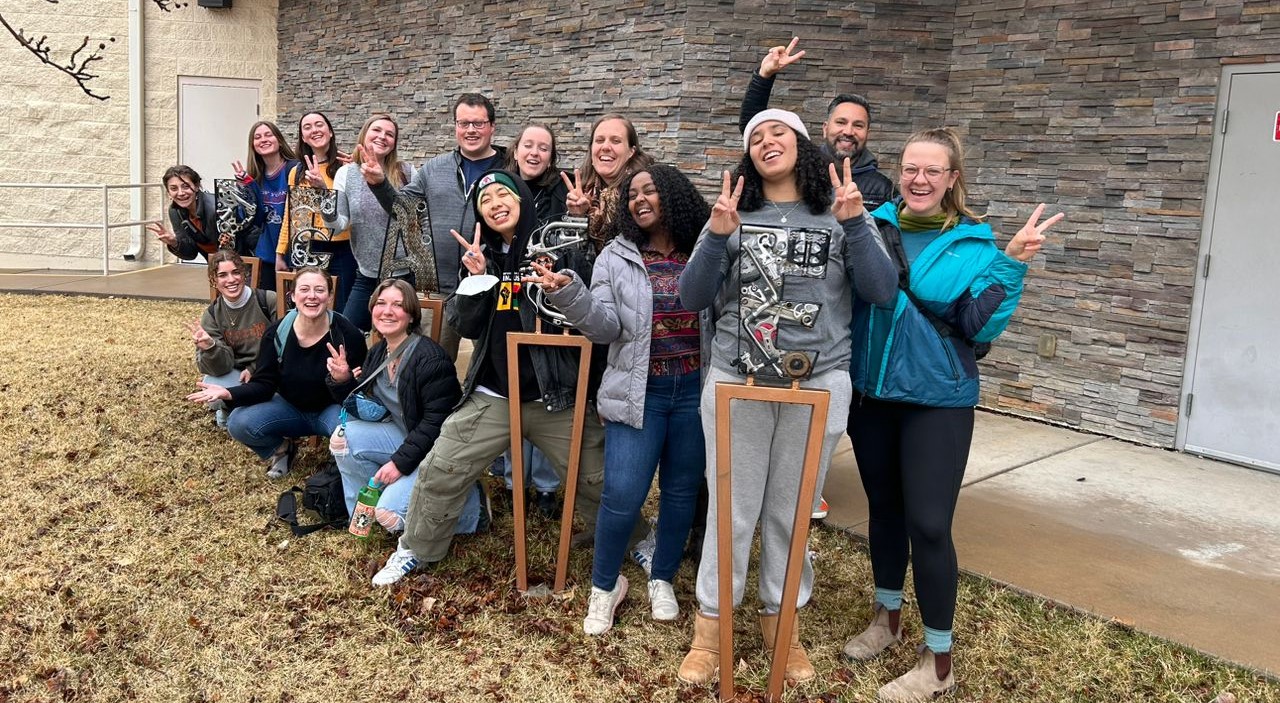 Group of PACS students and practitioners standing around yard signage made from recycled metals which spell out "P E A C E"