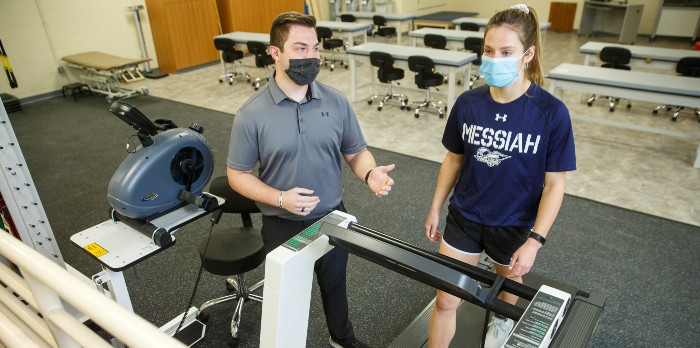 A man helps a student in an athletic training facility as she walks on a treadmill.