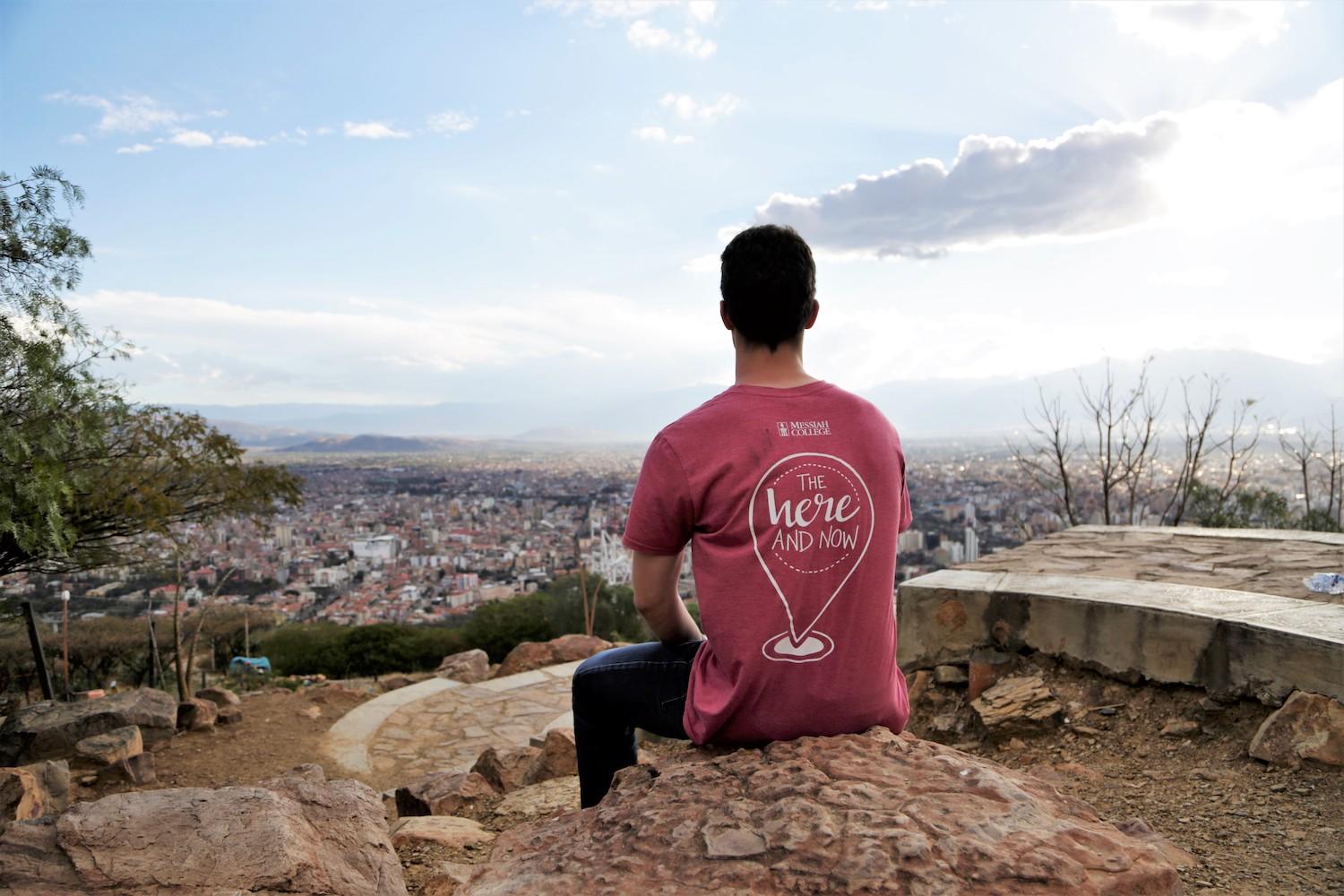 student sitting on a cliff looking at a city in the distance.