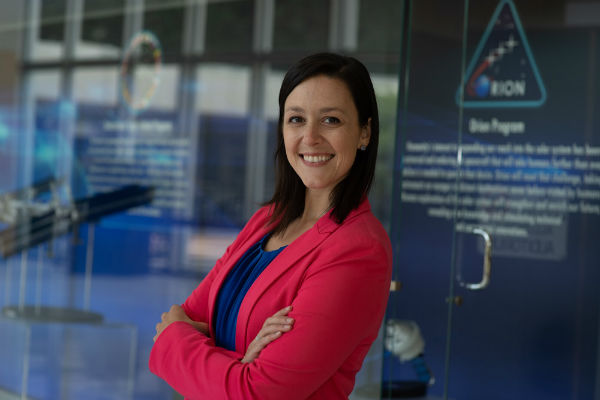 A woman stands with arms folded in front of glass doors smiling at the camera. The woman is wearing business clothing.