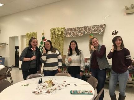 A group of students standing around a white table with chairs, holding up a tie, a red hat, a candy cane, a green hat and deer antlers (from left to right).