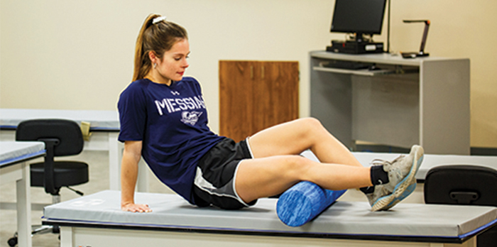 A woman in black athletic shorts and a blue Messiah shirt sits on a table with a foam roller under her leg.