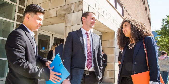 Two male and one female in business clothing stand talking in front of a building.