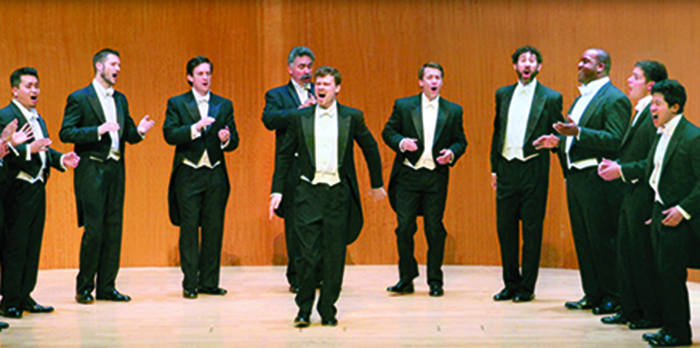 A group of men in tuxedos stand singing on a wooden stage.