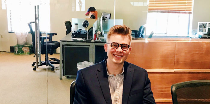 A male wearing a suit jacket and glasses sits smiling for the camera around a meeting table in a conference room.