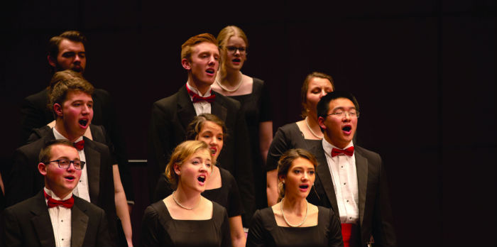 Students sing in a choir on stage in suits and dresses.